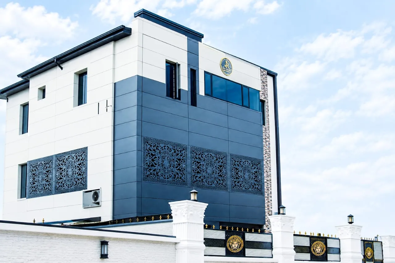 Corner of the Juls & Riff Lagos hotel: a three-storey building with dark grey and cream panels, carved decorative screens and a gold J&R emblem, behind a white perimeter wall