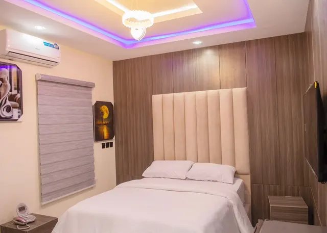 Mini Room with a tall cream headboard, taupe wood-panel wall, white bedding and a wall-mounted TV