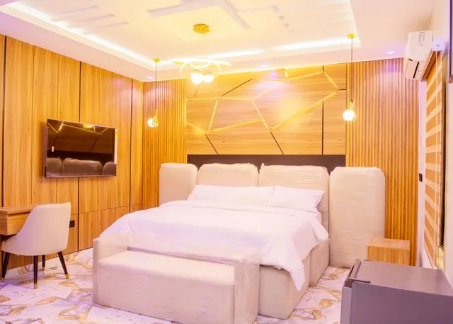 Royal Room with a lit geometric wood-panel headboard wall, a cream upholstered bed with bench, wall-mounted TV and desk